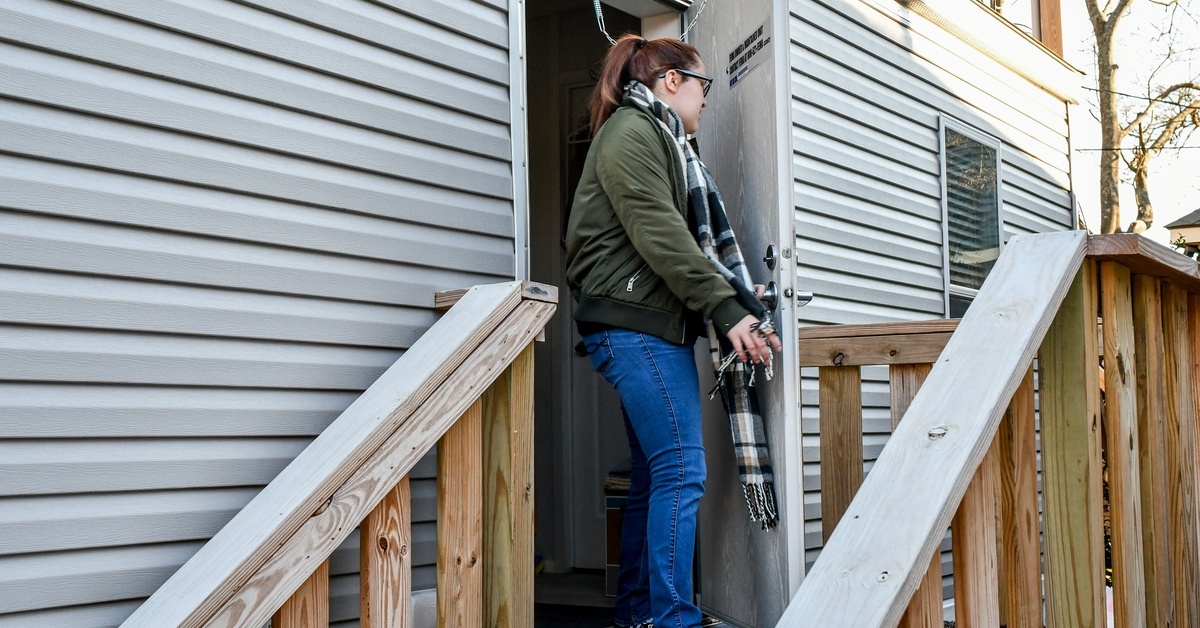 A young woman wearing a green jacket, jeans, and a black and white scarf is standing on a wooden porch. A young woman wearing a green jacket, jeans, and a black and white scarf is standing on a wooden porch.