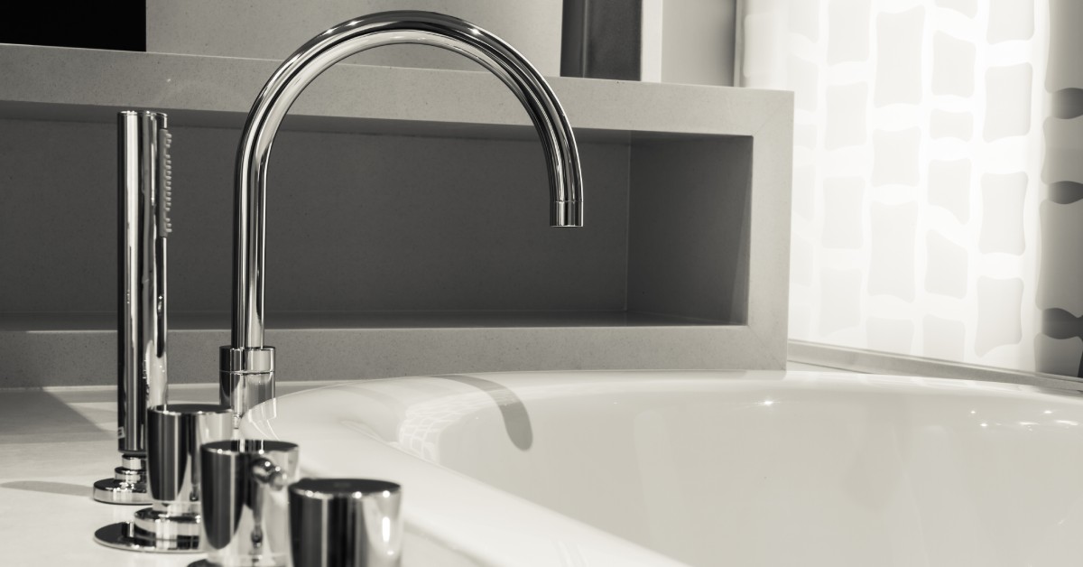 Light shines through a covered window near a white garden bathtub featuring a curved silver bath faucet.