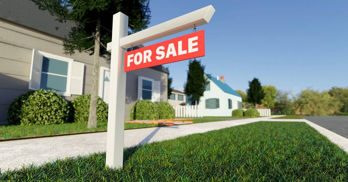 A real estate 'For Sale' sign is posted in the grass in front of a small home in a suburban neighborhood.