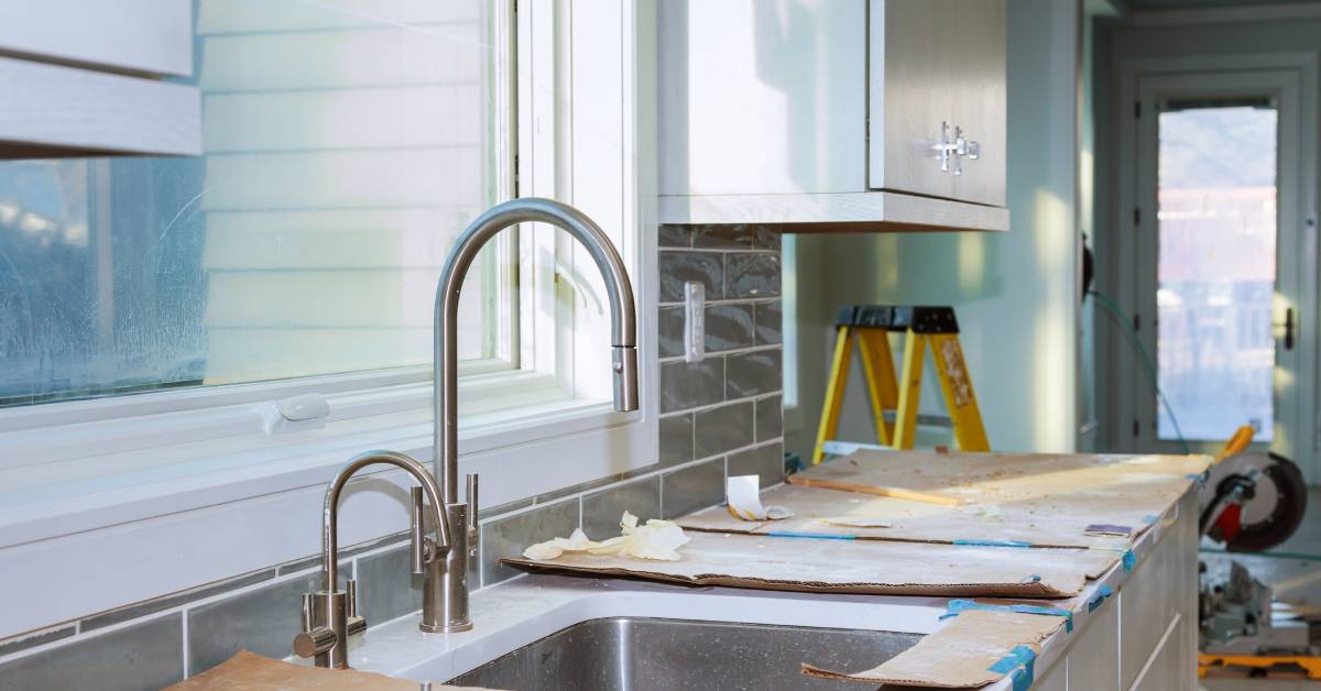 A kitchen in the midst of a remodel has white cabinets and a silver faucet. A ladder and a saw sit in the background.