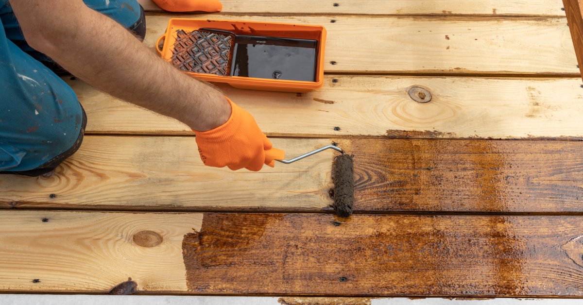 A person wearing orange work gloves kneels on a wooden deck and applies stain using a small paint roller.