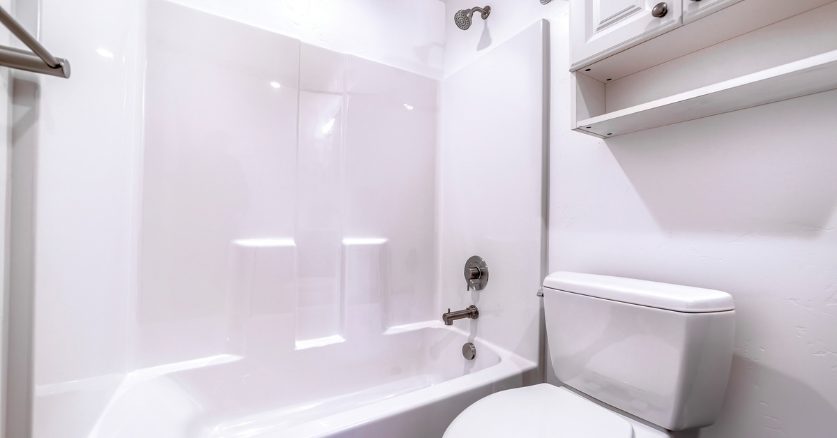 A bright white bathroom featuring a built-in bathtub, shower surround, and overhead storage above the toilet.