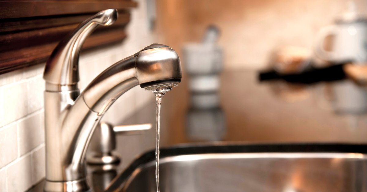 A silver kitchen faucet trickles water into a two-basin sink, set against a white tile backsplash and wooden window frame.