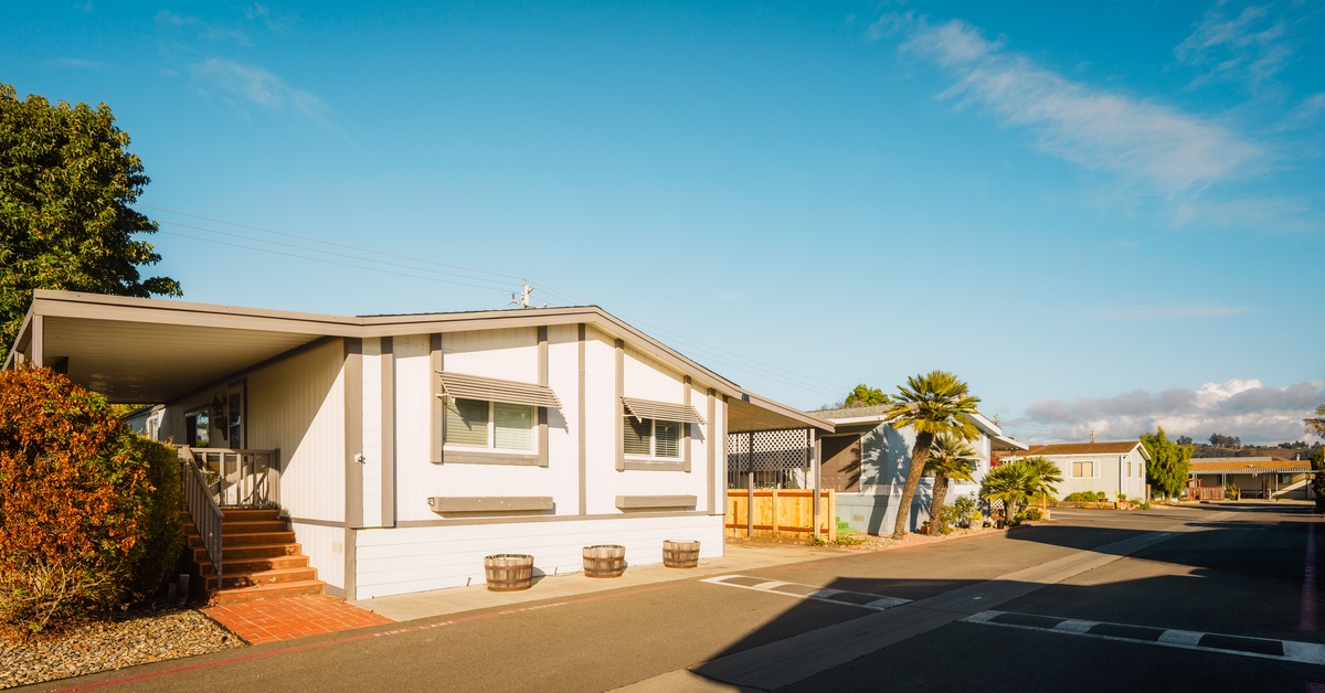 A row of light-toned mobile homes lines a neighborhood street under blue skies, with dry-climate plants in the landscape.