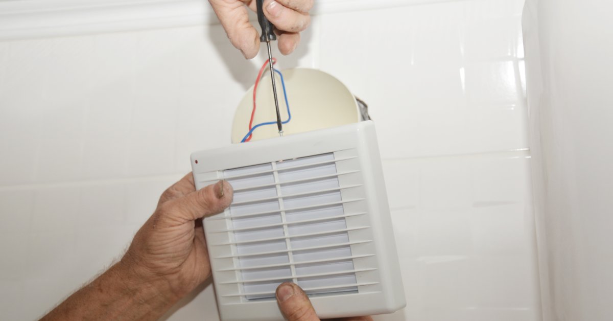 Two people, one holding the fan with both hands, one using the screwdriver, are replacing a bathroom exhaust fan.
