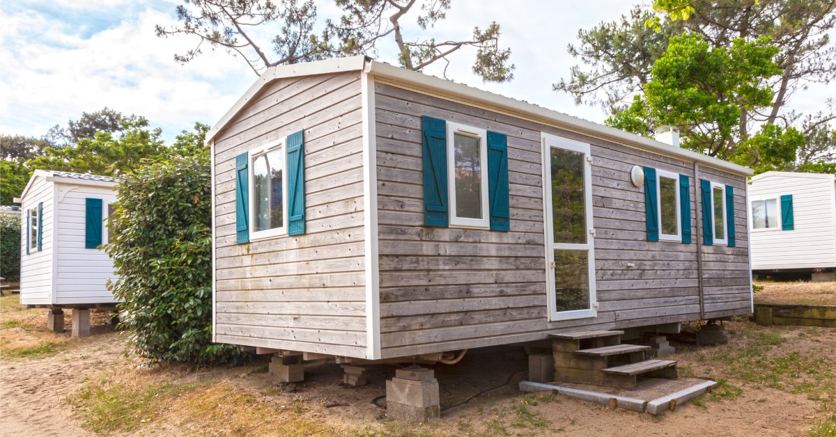 A mobile home with worn wood siding features teal shutters, a mid-view storm door, and stairs leading to the front entrance.