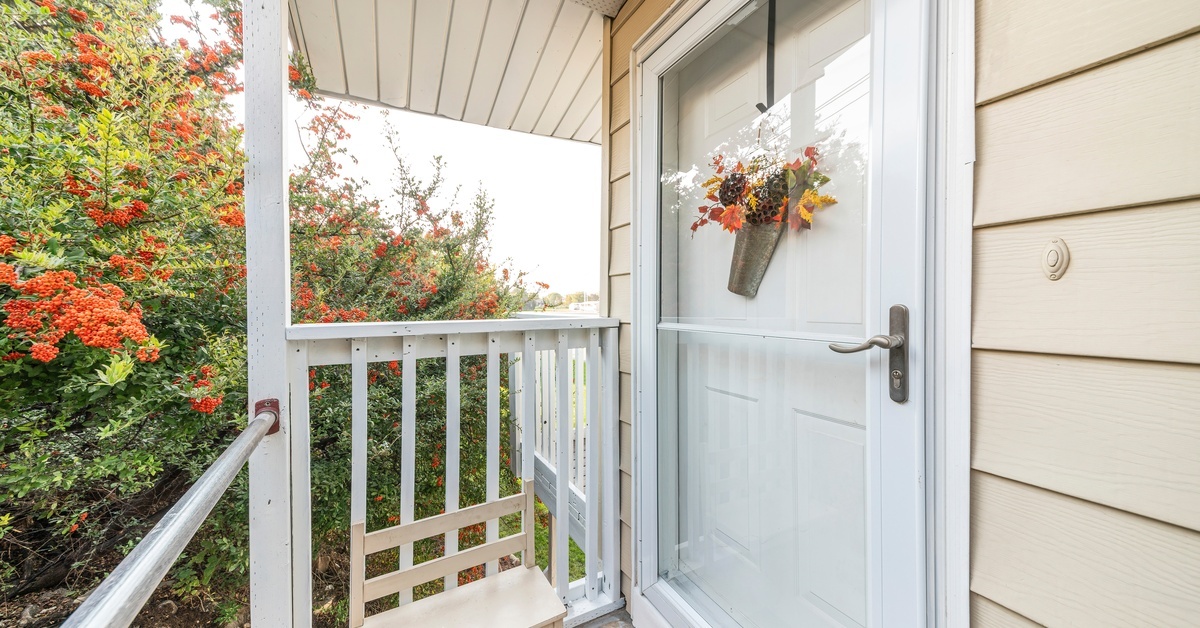 A front porch with tan siding features a white door with a glass storm door, and flowers hanging on the door. A front porch with tan siding features a white door with a glass storm door, and flowers hanging on the door.