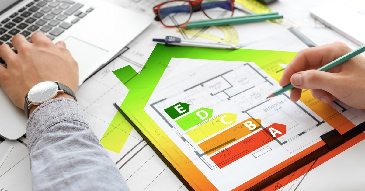 A person works at a laptop while holding a pencil, and a holographic energy efficiency design overlays some house plans.
