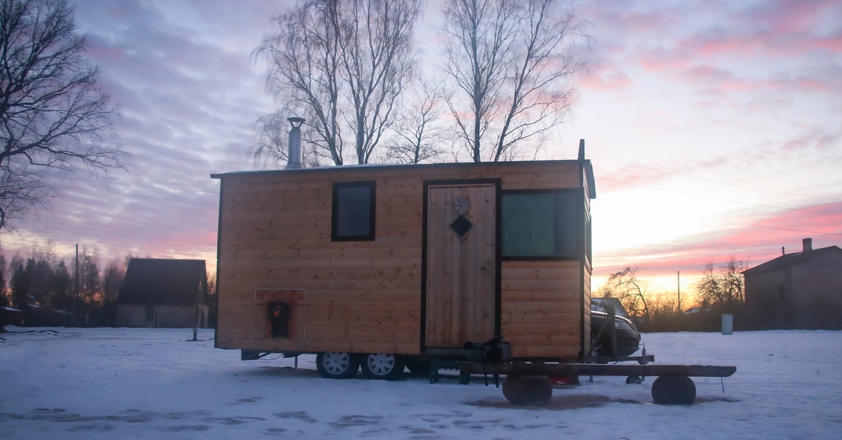 A small mobile home with wood siding and black trim sits in a snowy yard with the sun near the horizon behind it.