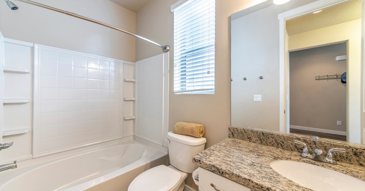 A home bathroom with a tub and shower, a toilet beneath a window, and a sink with granite counters and white cabinets.