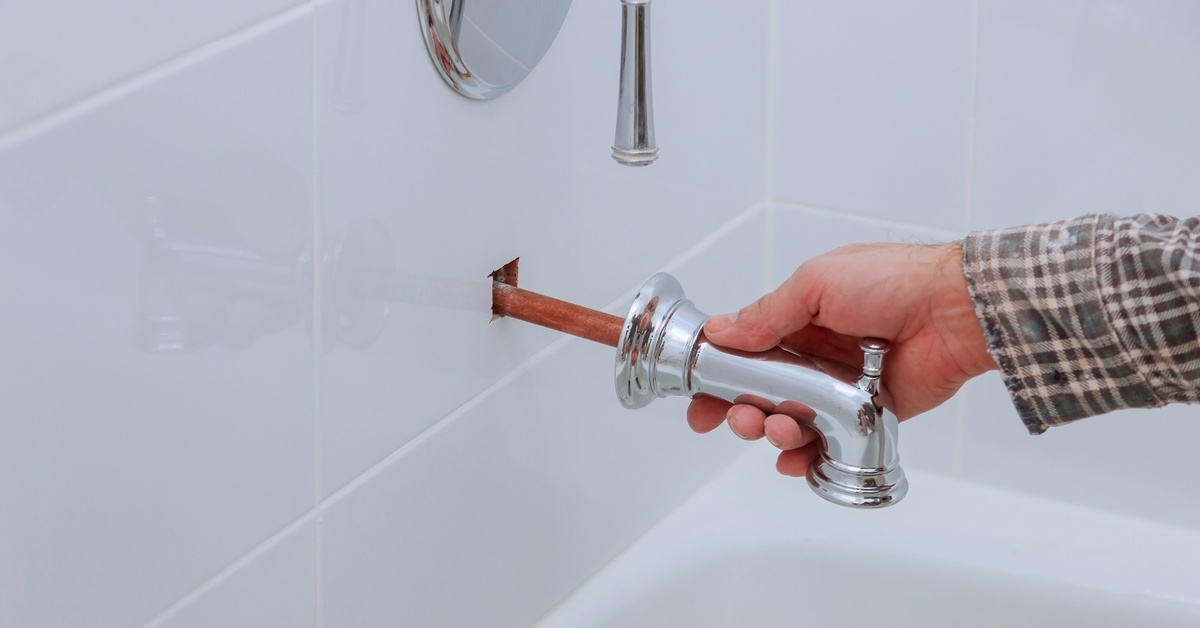 A person installs a tub water faucet into a white tiled wall underneath a matching temperature dial.