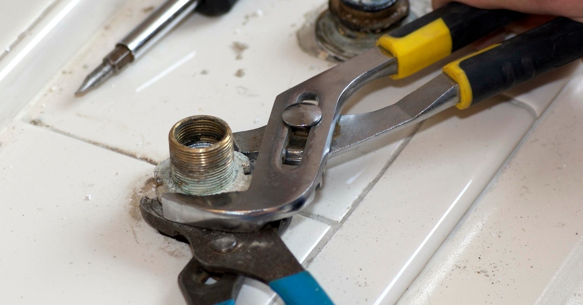 A person uses two wrenches to loosen a water connection set in tile, with a screwdriver lying nearby.