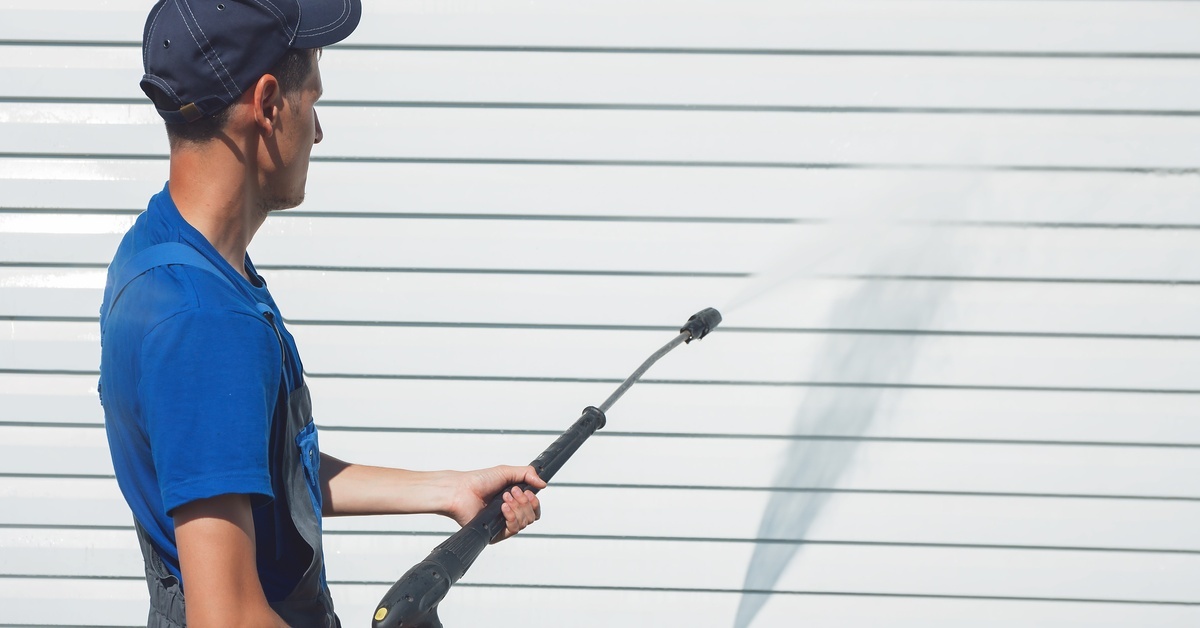 A man wearing waterproof coveralls using a power washer to clean the white siding of a house on a sunny day.