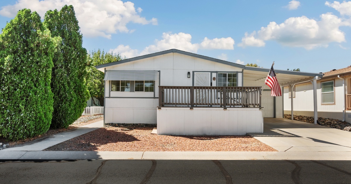 A white double-sided mobile home that has a porch, garage, and carport, with an American flag hanging out front.