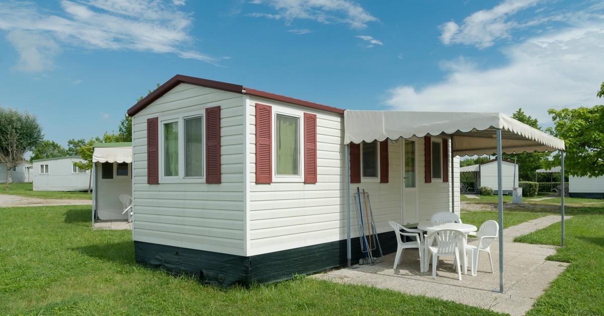 A small white trailer home with red shutters sits on green grass. A white awning shades chairs on a concrete pad out front. A small white trailer home with red shutters sits on green grass. A white awning shades chairs on a concrete pad out front.