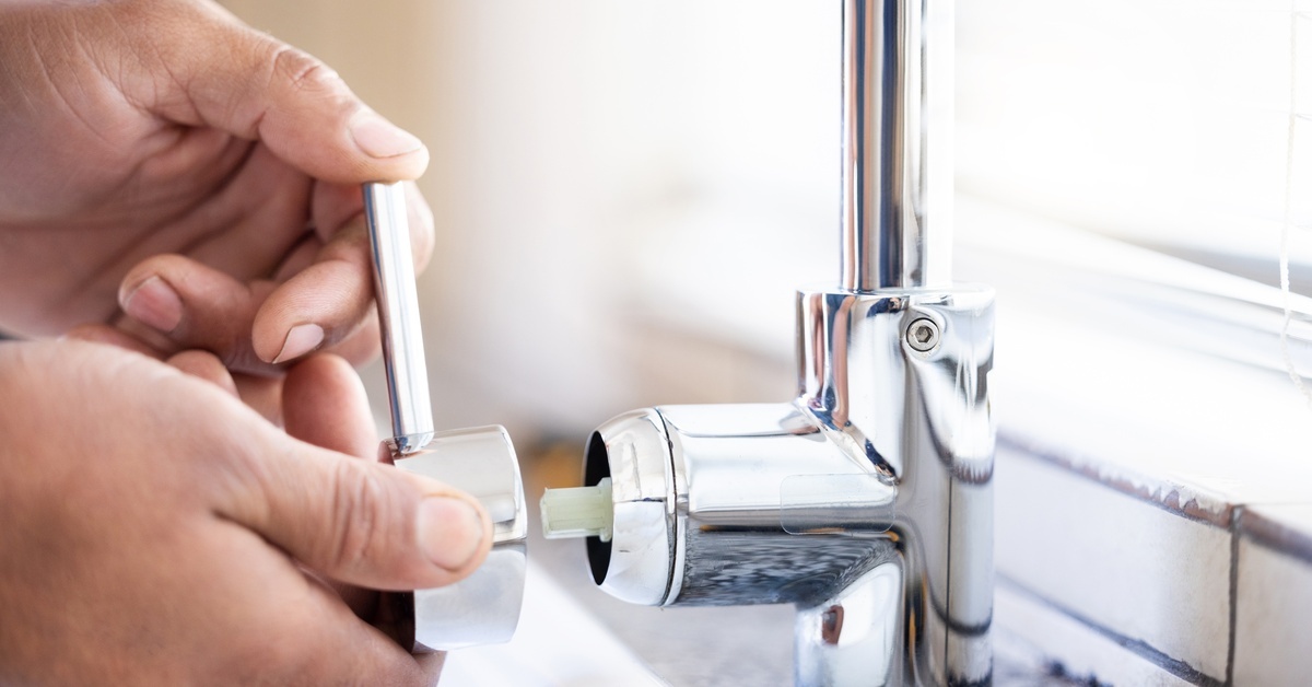A person’s hands gripping a handle near a silver faucet to make a repair while sunlight shines in through a window.