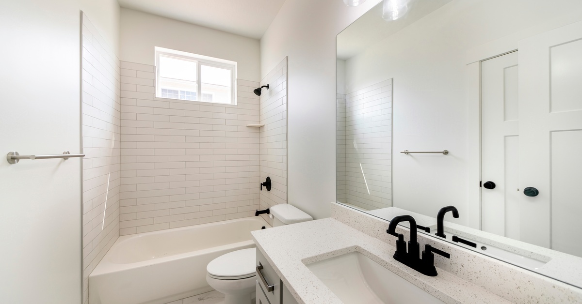 An empty bathroom featuring white walls, a tub, tiles, counters, and flooring, with gray cabinets and black fixtures.