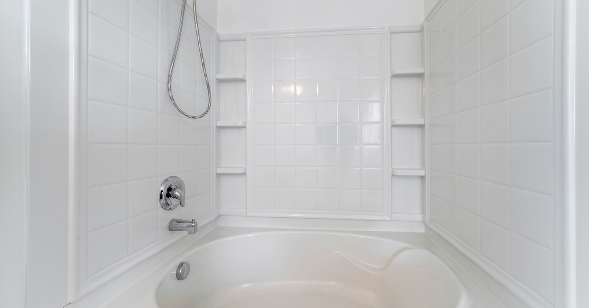 A white tub and shower combo with a chrome handheld shower fixture, wall-mounted faucet, and a white tile-look surrounding the tub.