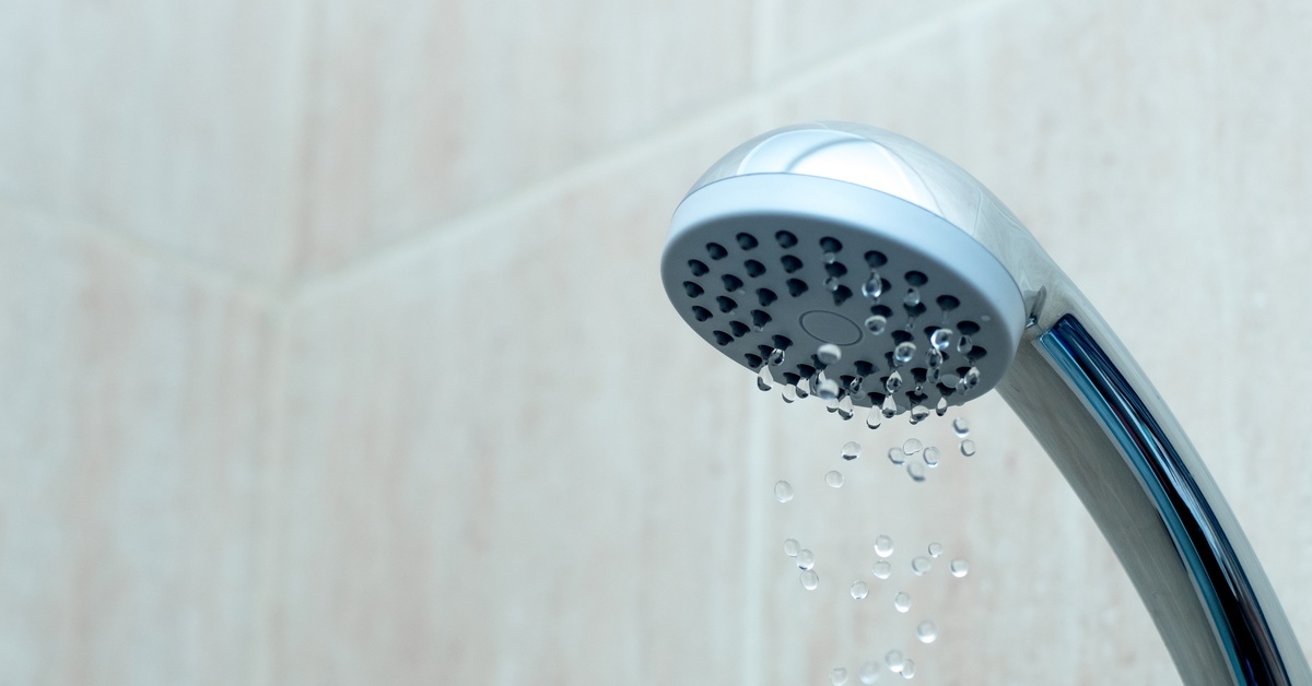 A close-up of clean water trickling weakly out of a silver showerhead in a bathroom with tiled walls.