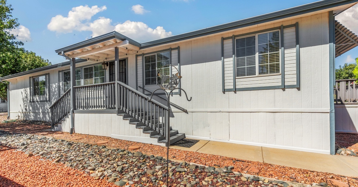 A gray mobile home with dark trim has a double stair porch, a concrete sidewalk, and red and gray stone landscaping.