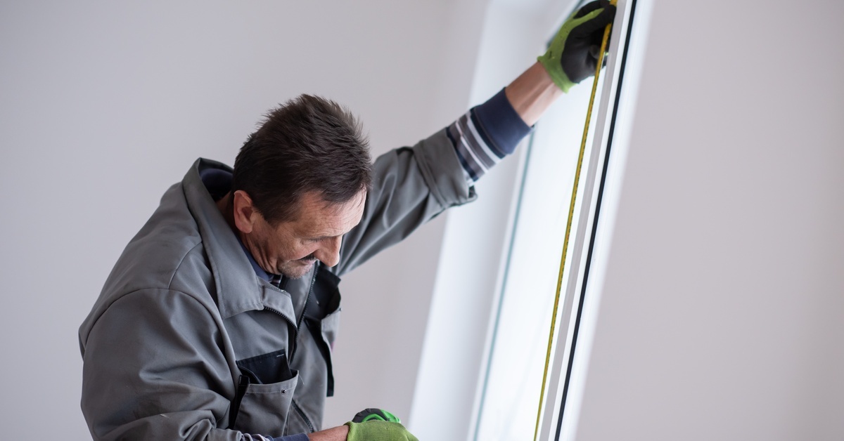An older man in work gear uses a measuring tape on the right side of a window inside a home with white walls.