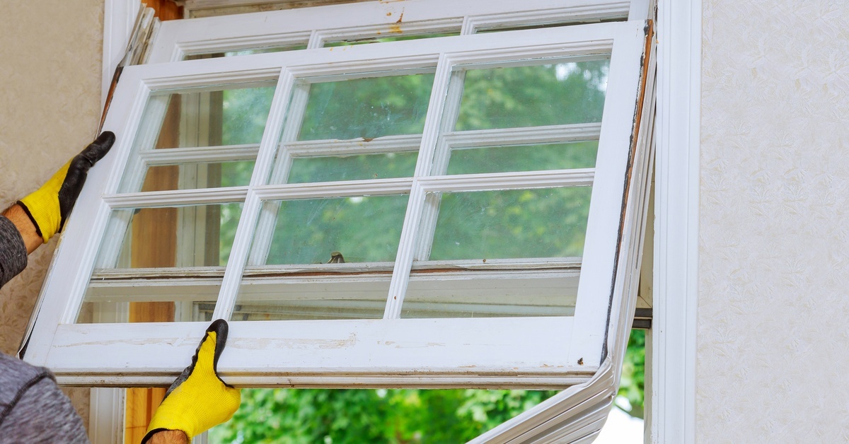 A person wearing yellow and black gloves pulls an old window inward from a wall opening inside a house.