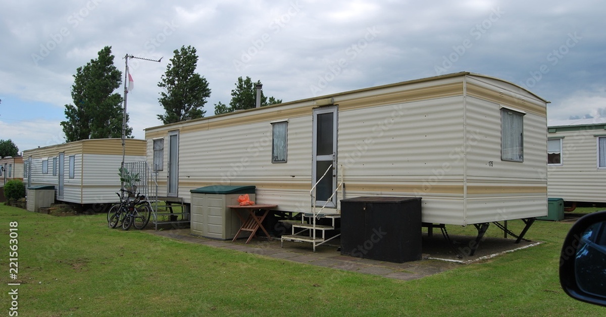 A tan mobile home with green grass and no skirting outside. There are storage units near the front doors and other mobile homes surrounding the area.