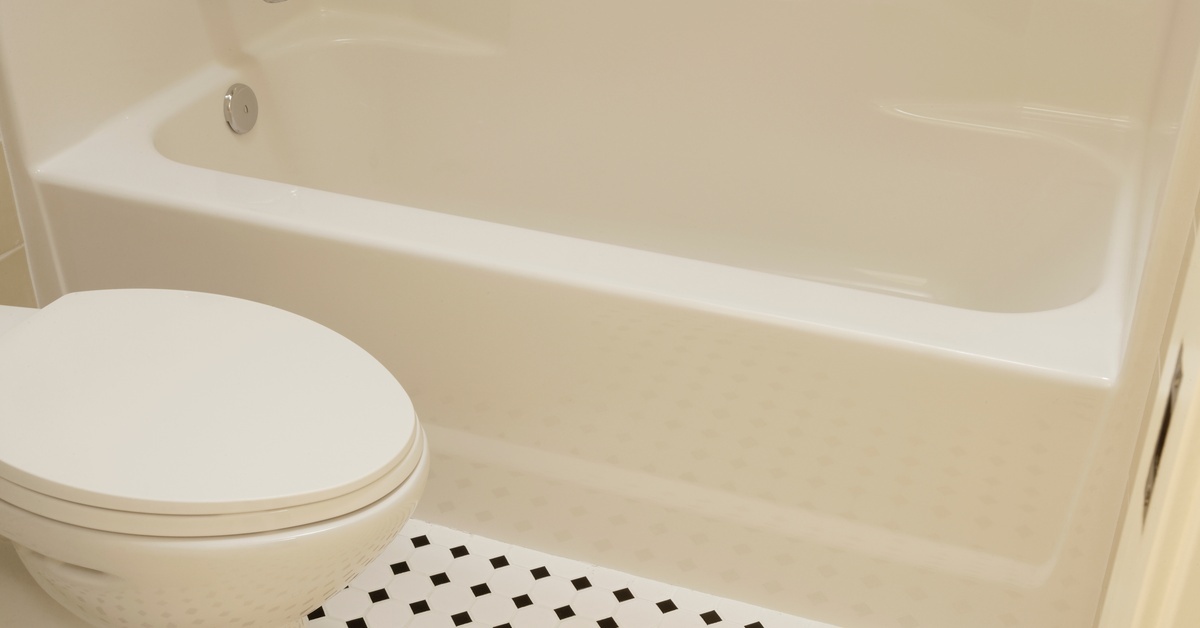 A white toilet is next to a fiberglass bathtub with chrome fixtures. The flooring is made of black and white diamond tile.