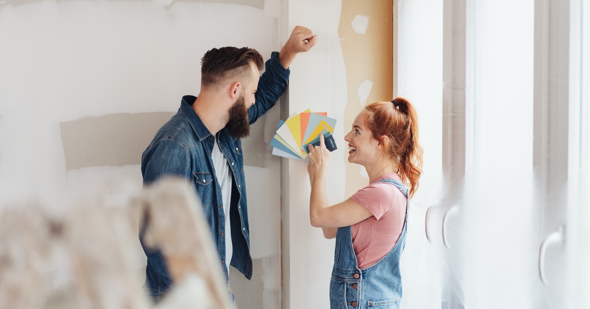 A man in a denim shirt and a woman in overalls smile at each other while looking at paint swatches in a room to be painted.