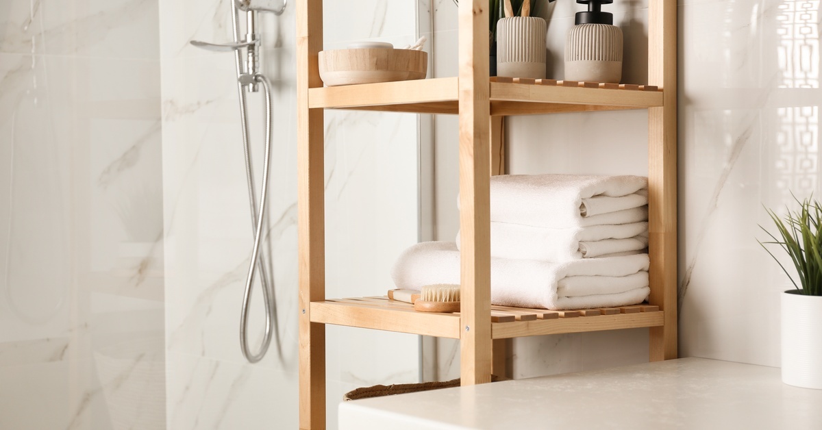 A light wood open shelving unit stands against a stone wall in a bathroom with white countertops and chrome fixtures.
