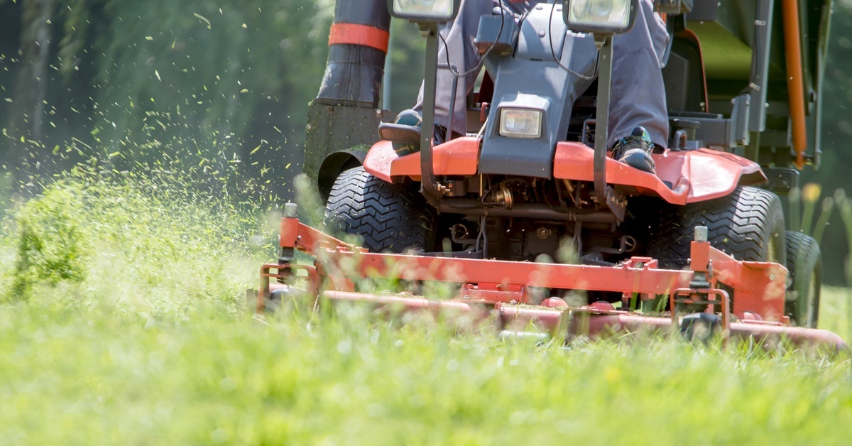 A person on a red riding lawnmower cuts bright green grass, with clippings flying to the side as the mower moves forward.