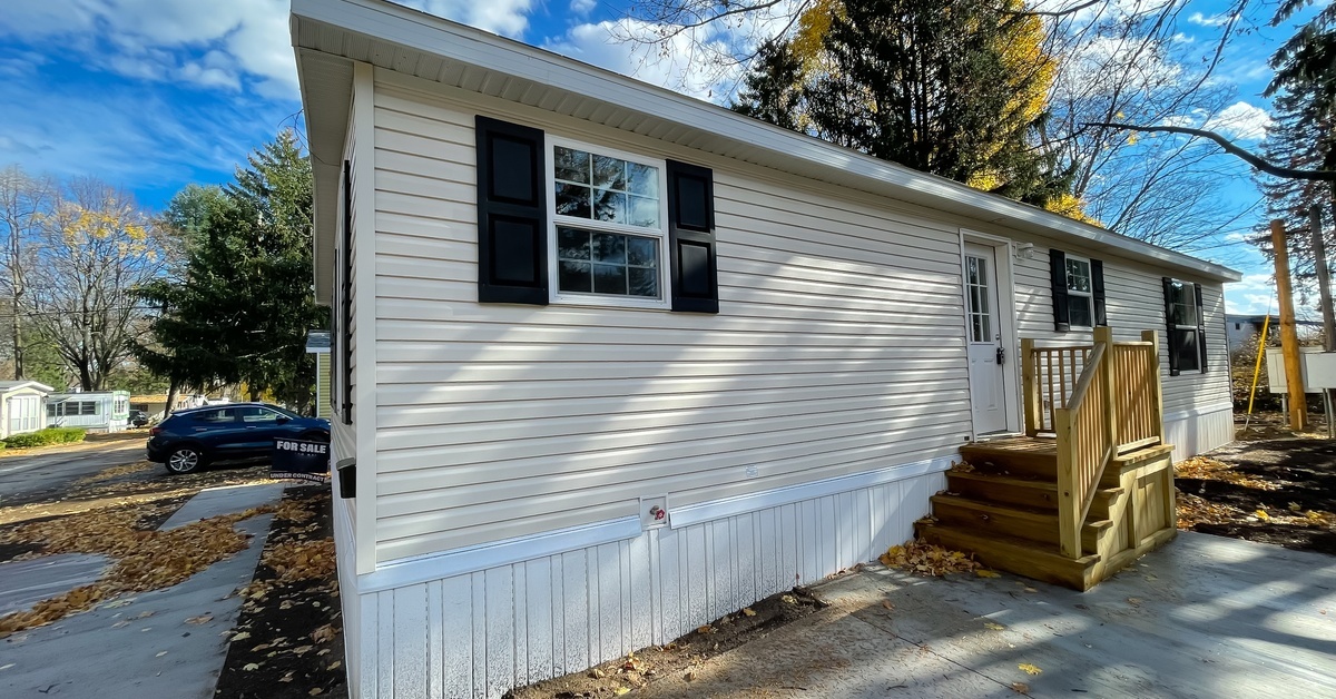 A tan mobile home with white skirting and black shutters has a small wood staircase and fallen autumn leaves around it.