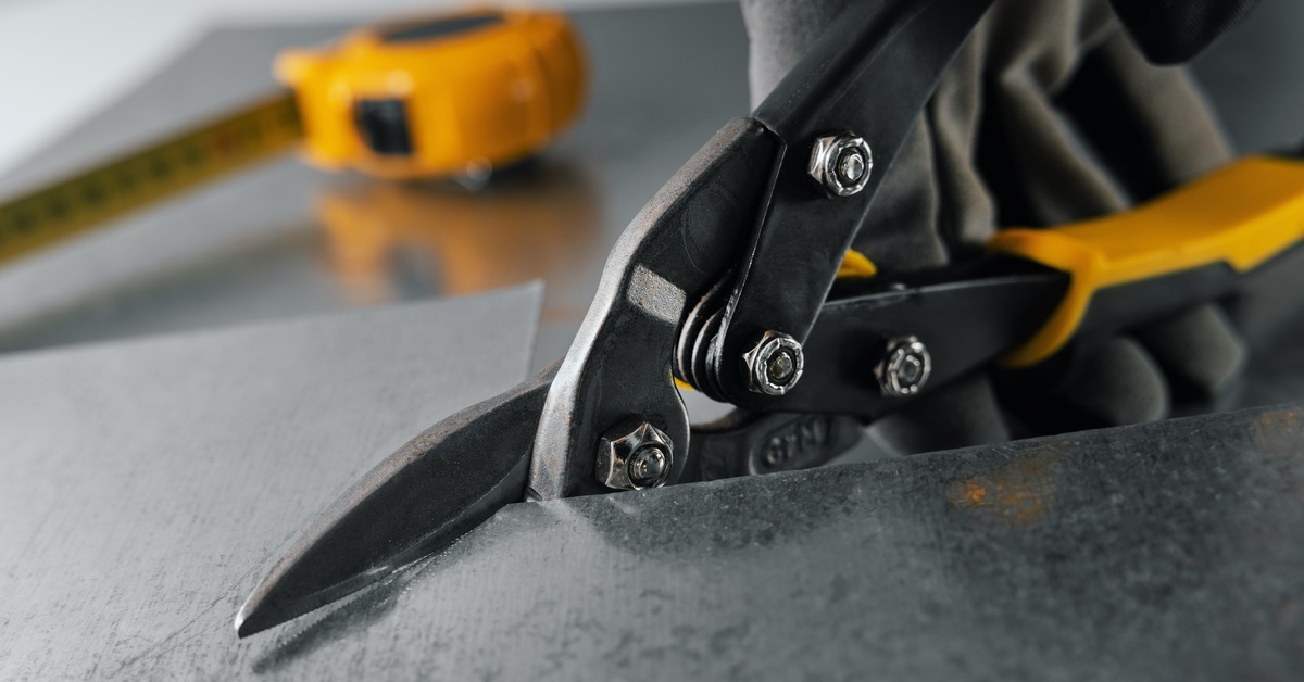 Someone in black gloves uses yellow tin snips to cut thin metal, with a measuring tape resting on another metal piece nearby.