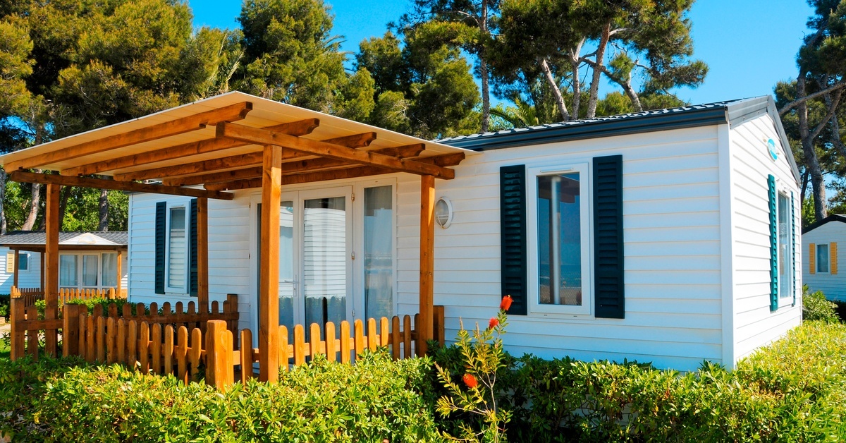 A mobile home features a front porch with a wooden fence and roof. Bushes line the edges of the porch and home.