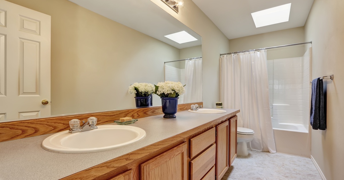 A long bathroom in a mobile home with stained wood vanity cabinets and a large mirror. There's a surround tub shower unit.