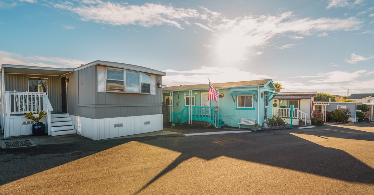 The sun beams down on two mobile homes with white and turquoise skirting panels in a residential mobile home park.