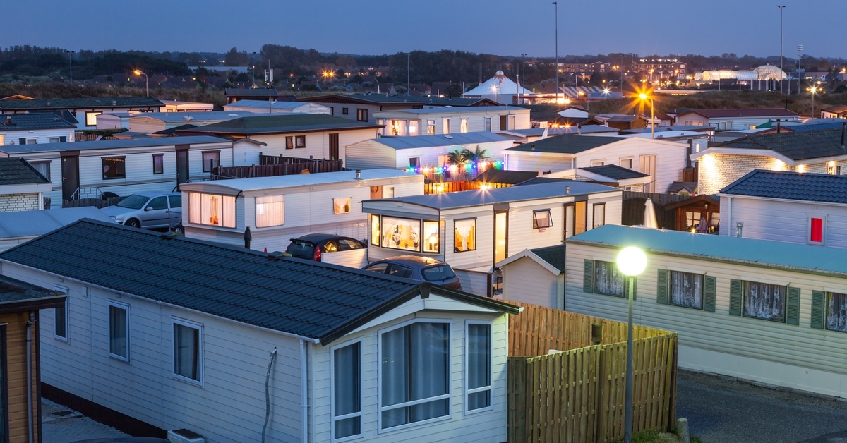 A manufactured home community at dusk with closely spaced homes, lit windows, parked cars, and streetlights glowing at night.