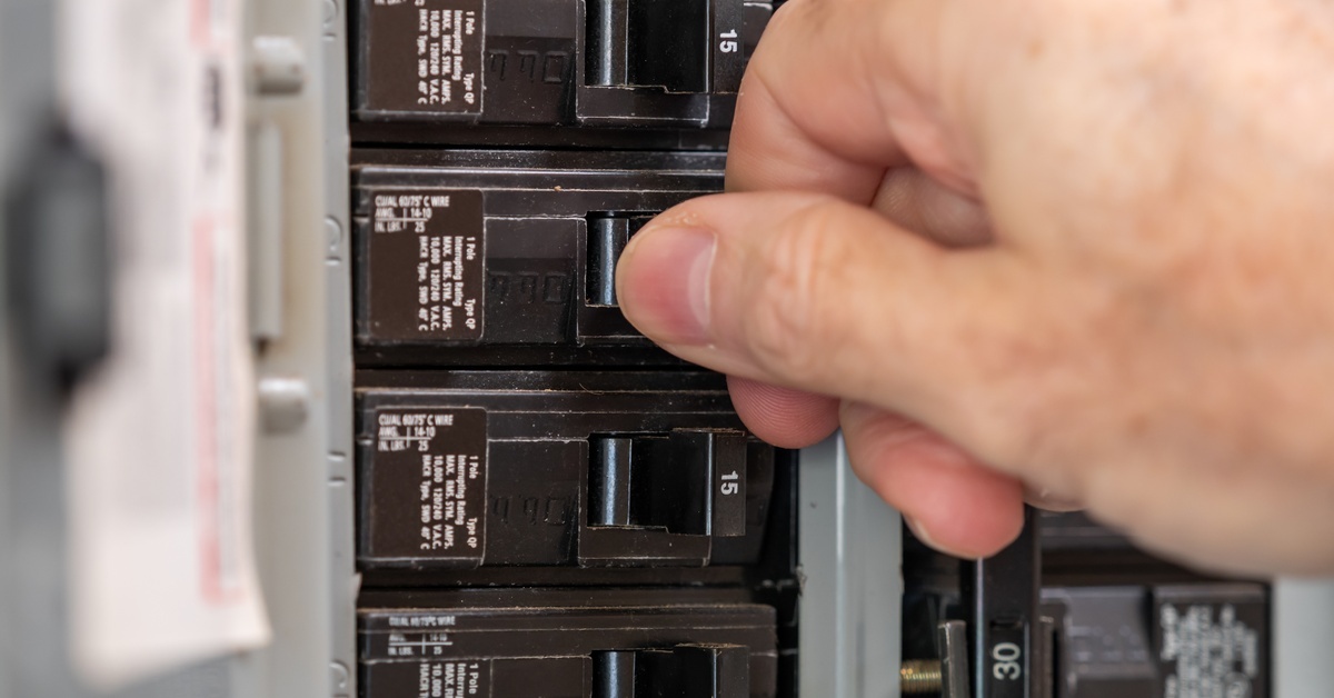 A close-up of a hand switching off a breaker in an open-circuit breaker box. A breaker map is blurred inside the door.