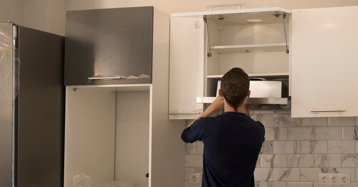 A handyman wearing a black shirt installs a range hood onto white cabinetry above a stove in a new kitchen.