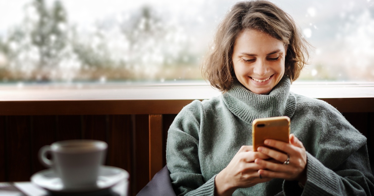 A woman in a cozy gray sweater smiles as she looks at her phone by a window overlooking a snowy winter landscape. A woman in a cozy gray sweater smiles as she looks at her phone by a window overlooking a snowy winter landscape.
