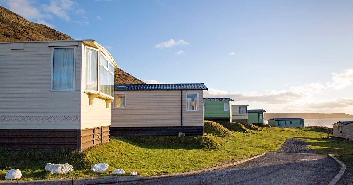 A row of mobile homes on a grassy hillside with visible skirting underneath, parked along a curved driveway under a blue sky. A row of mobile homes on a grassy hillside with visible skirting underneath, parked along a curved driveway under a blue sky.