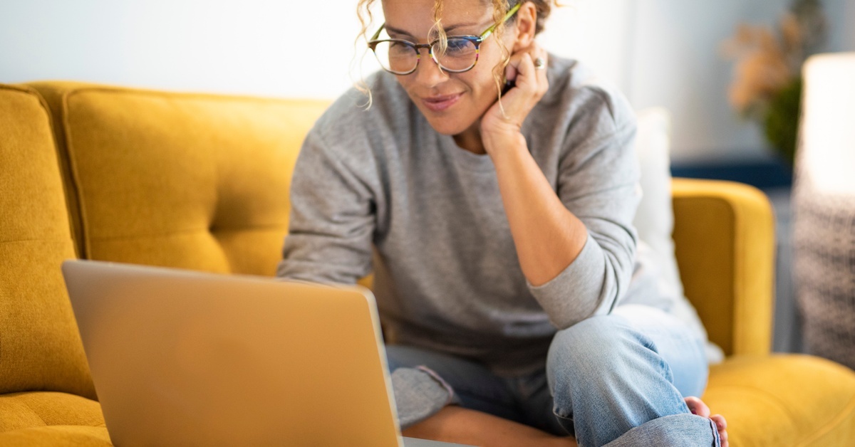 A woman wearing a grey sweater, glasses, and a messy hairdo sits on a yellow couch, staring intently at her laptop.
